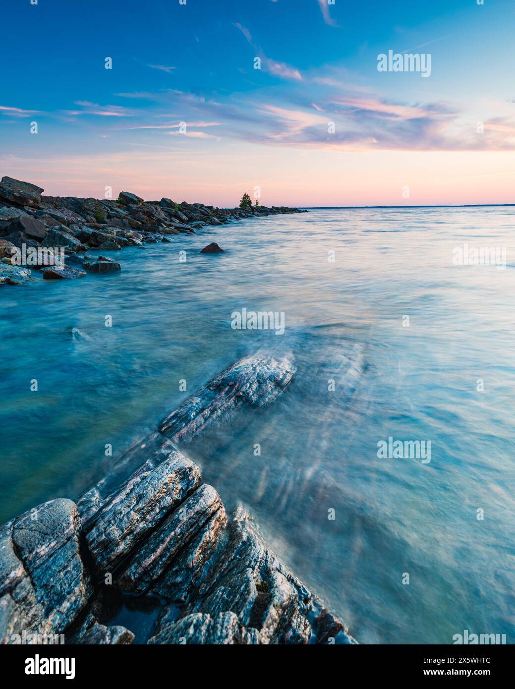 A body of water, Lake Vänern, is seen at sunset, bordered by a rocky shore. The calm waters meet the rugged rocks in a picturesque scene. Stock Photo