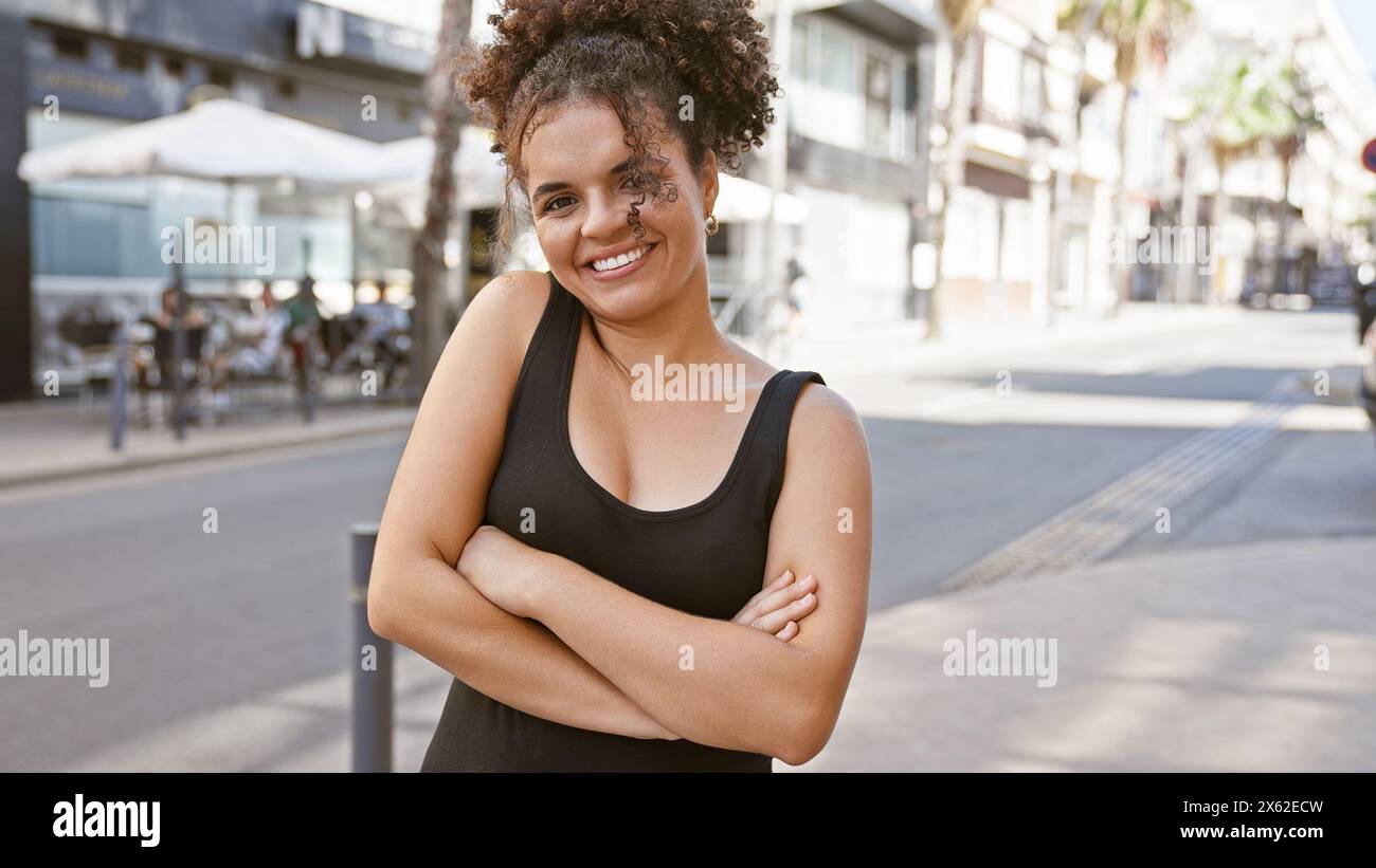 Smiling young adult hispanic woman with curly hair standing crossarmed on a sunny urban street. Stock Photo