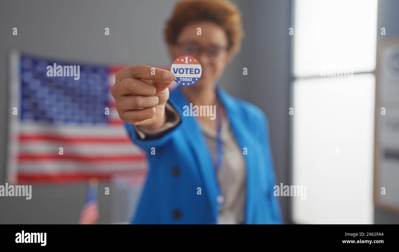 African american woman in a blue blazer showing 'i voted' sticker with us flag background indoors Stock Photo