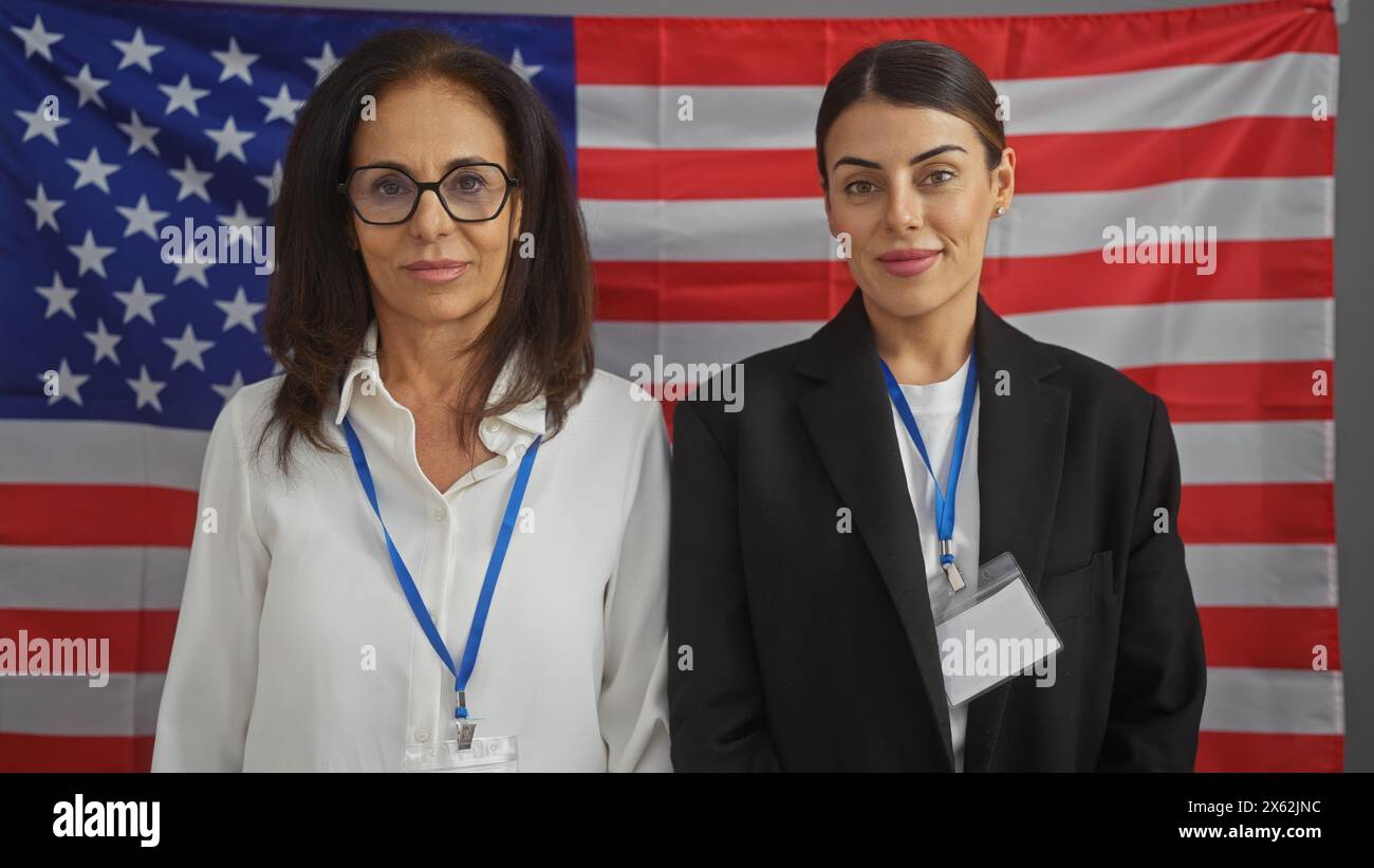 Two professional women with id badges stand confidently in an american office, backed by a usa flag. Stock Photo