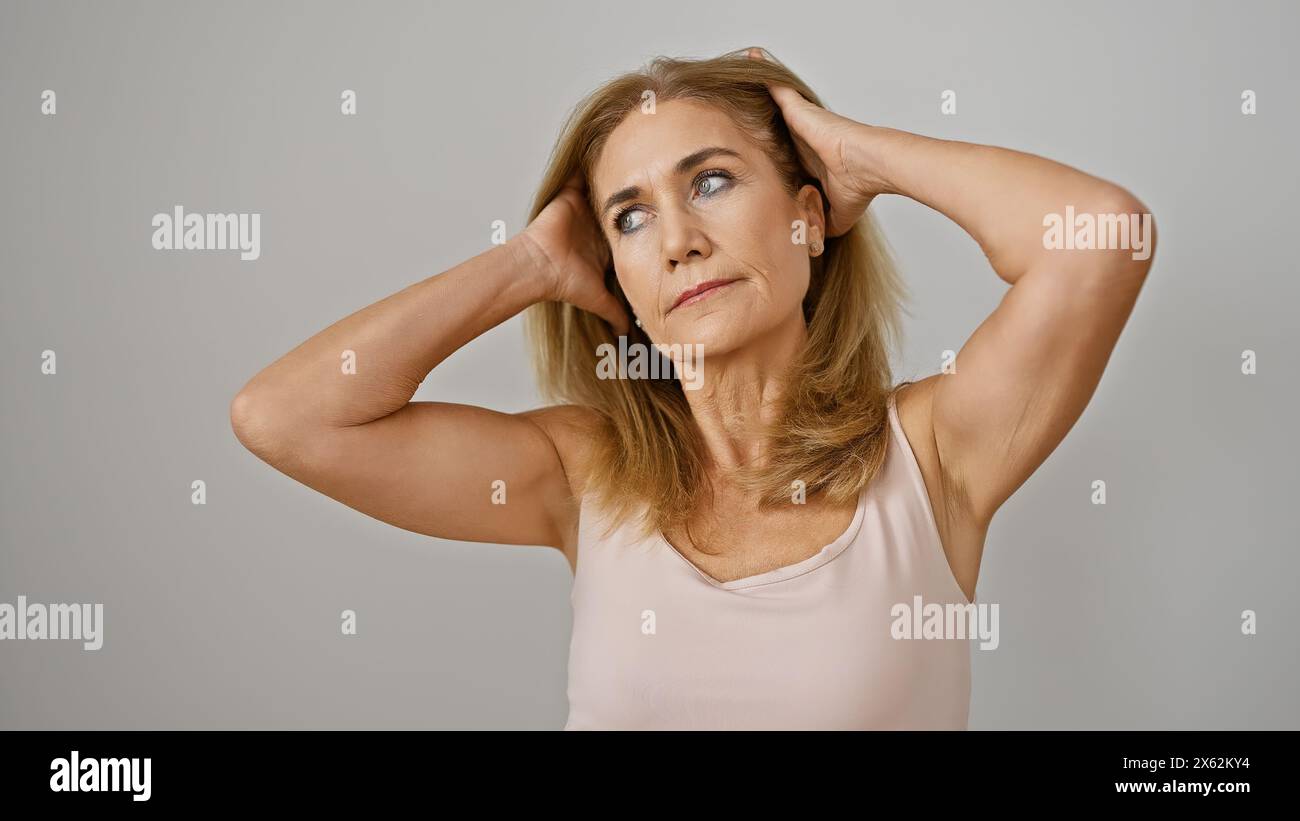 A contemplative blonde woman in a sleeveless top isolated against a white background poses with her hands behind her head. Stock Photo