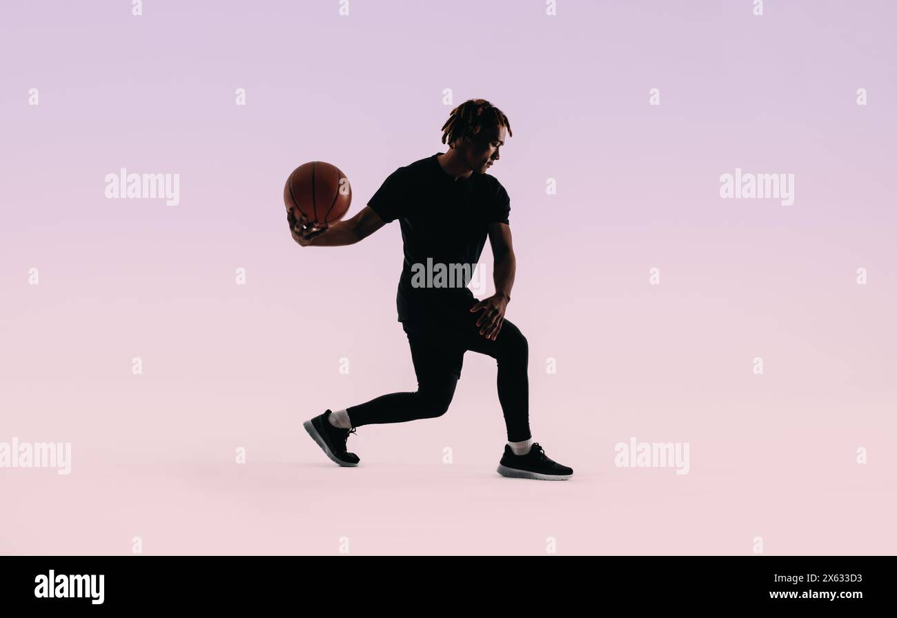 Silhouetted against a basketball court background, a young athlete with dreadlocks practices dribbling a ball with perfect form. He wears sportswear a Stock Photo