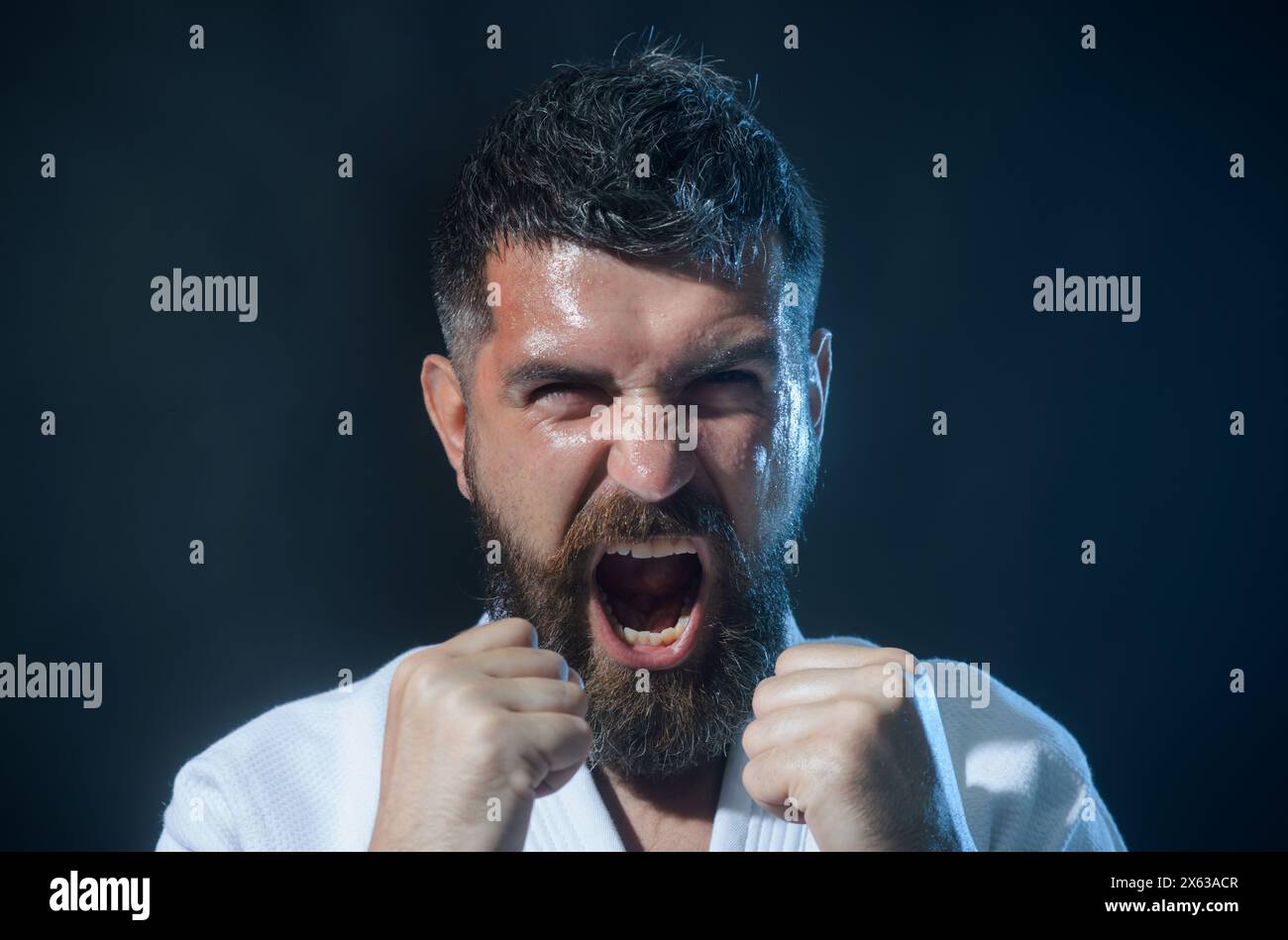 Angry screaming bearded man in white kimono in fighting position. Karate fighter preparing for competition. Muscular male fitness model ready for Stock Photo
