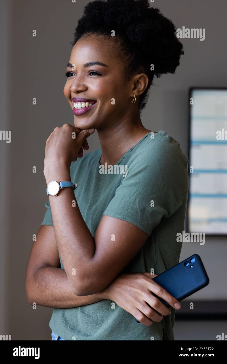 At office, African American businesswoman holding smartphone, looking away, smiling Stock Photo