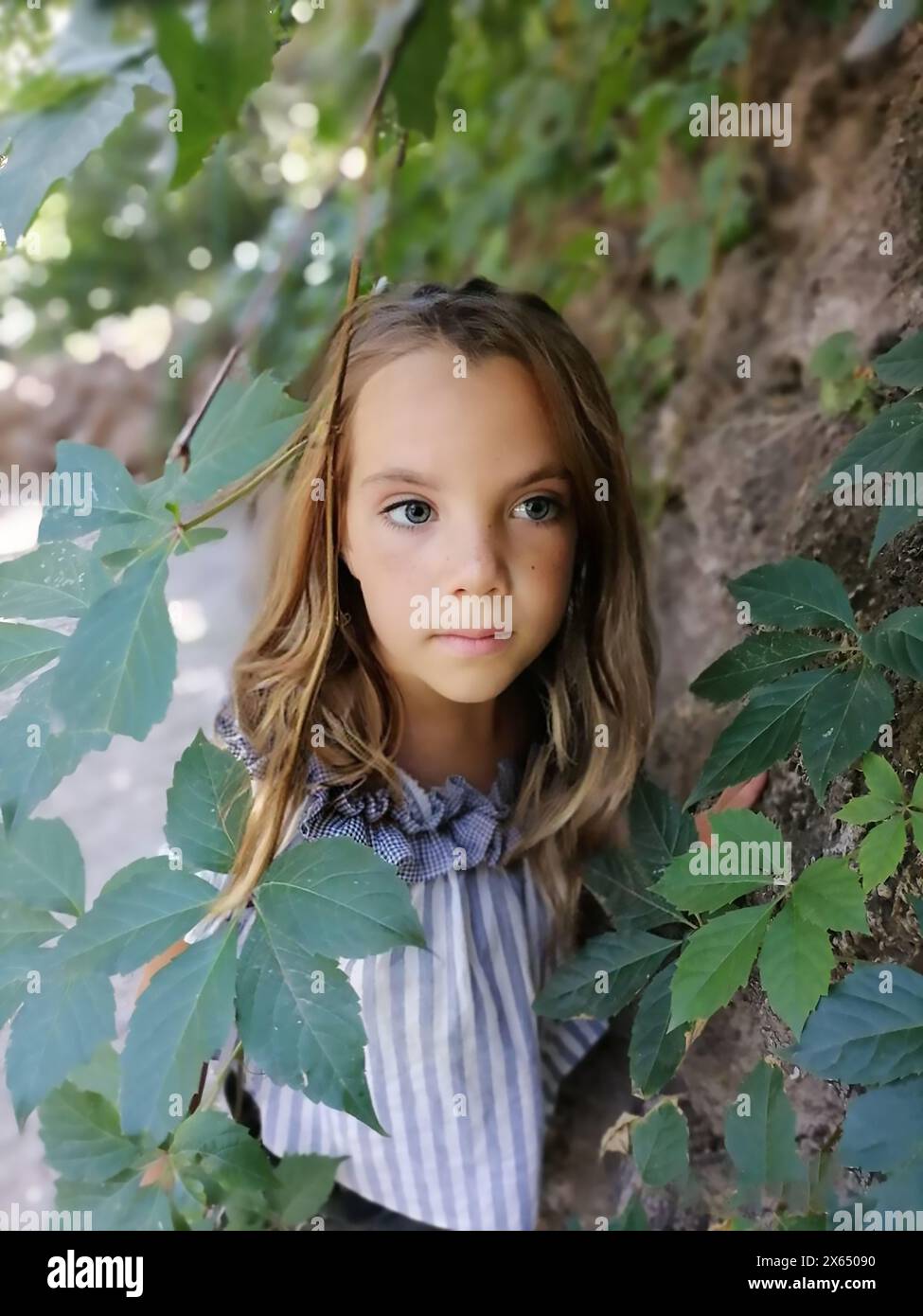 Little girl in the garden. Beautiful flowers roses. Cute lady watching thoughtfully. Child with big green eyes. Lovely flowers pink color. Green plant Stock Photo