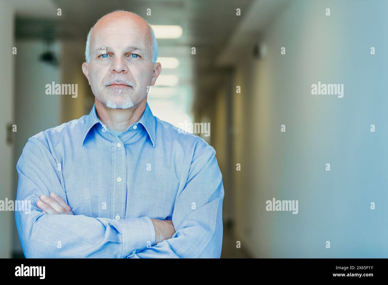 Mature man exuding confidence in a blue shirt, arms crossed, in front of a window showing blurred greenery Stock Photo