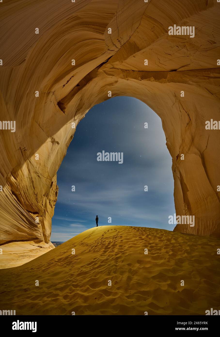 A lone hiker stands on a sand dune, dwarfed by a massive alcove Stock Photo
