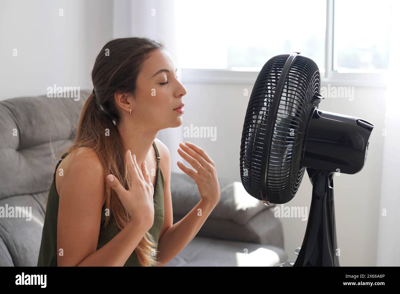 Summer heat. Young Hispanic woman cooling down by ventilator at home, feeling unwell with high temperature during hot weather. Latin girl in front of Stock Photo