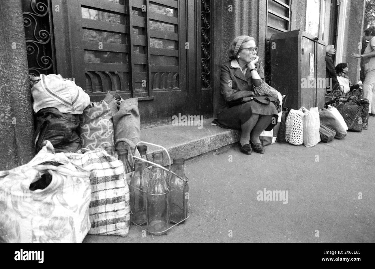 Bucharest, Romania, February 1990. Shortly after the anti-communist revolution of December 1989, people still stay in long lines to get hold of basic groceries. The centralized socialist economic system created scarcity and hunger. Here, an elderly woman is sitting down, waiting in line for the delivery of milk. Other people left their bags with reusable glass bottles, reserving their spot in line. Stock Photo