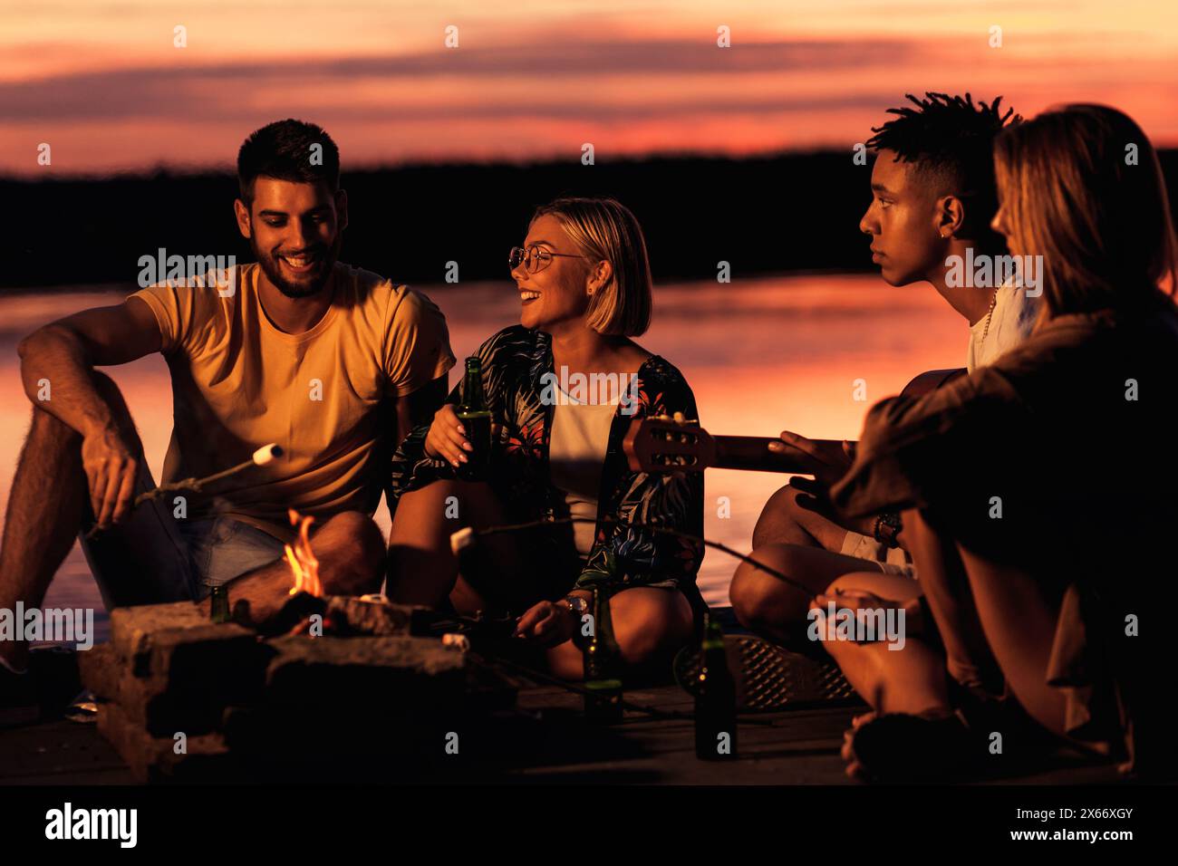 Group of young friends enjoying at the lake at night. They sitting around the fire singing and having fun. Stock Photo