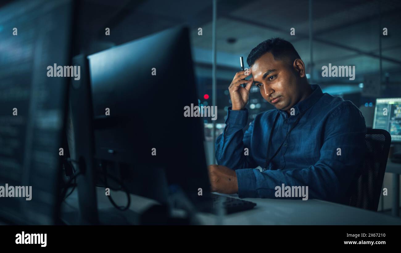 Night Office: Portrait of Handsome Indian Man in Working on Desktop Computer. Digital Entrepreneur Typing, Creating Software, e-Commerce App Design, Programming. Thoughtful Man Seeking Solution Stock Photo