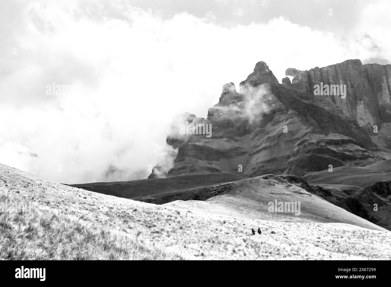 Distant hikers walking towards the ominous, mist covered peaks of the Drakensberg Mountains of South Africa in black and white Stock Photo