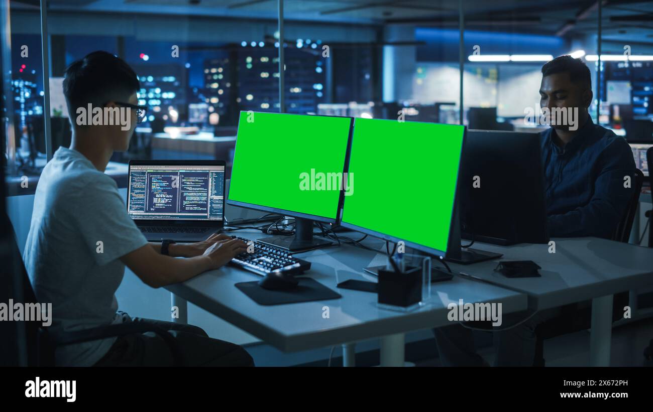 Night Office: Young Japanese Man in Working on Green Screen Chroma Key Desktop Computer. Team of Programmers Typing Code, Creating Modern Software, e-Commerce App Design, e-Business Programming Stock Photo