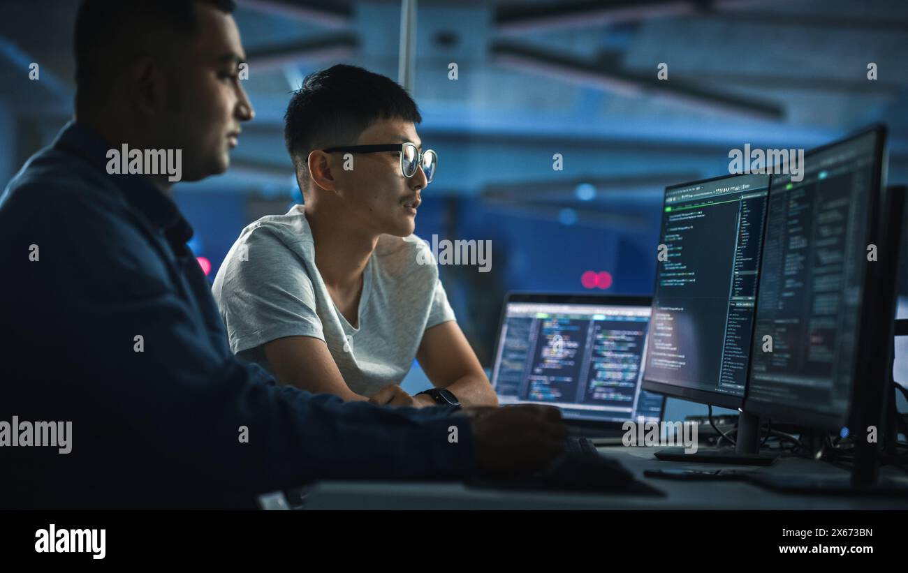 Teamwork In Diverse Office at Night: Project Manager Talks with Creative Solutions Specialist, They Discuss App Code Computer. Stylish Professionals Programming, Create e-Commerce Software Stock Photo
