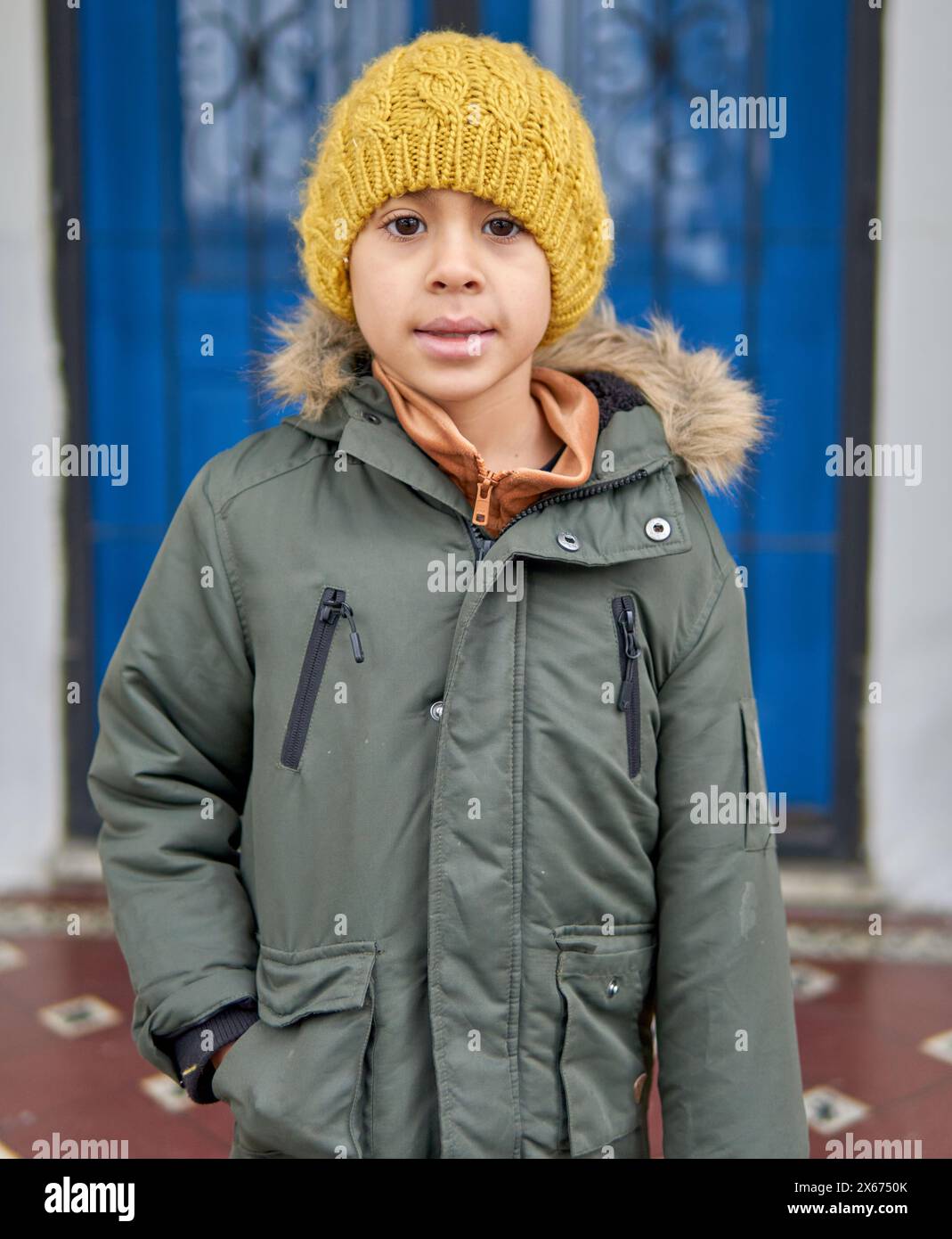 latin boy standing facing the camera with a warm jacket and wool cap, in the background a light blue door in Tucuman, Argentina. Stock Photo