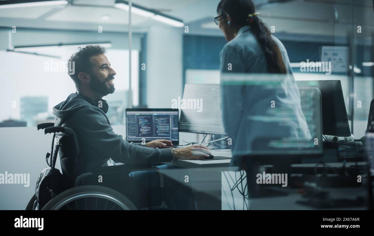 Disability-Friendly Office: Brilliant IT Programmer with Disability in a Wheelchair Talks with Colleague while Working on Computer. Software Engineers Develop Innovative App, Program. Stock Photo