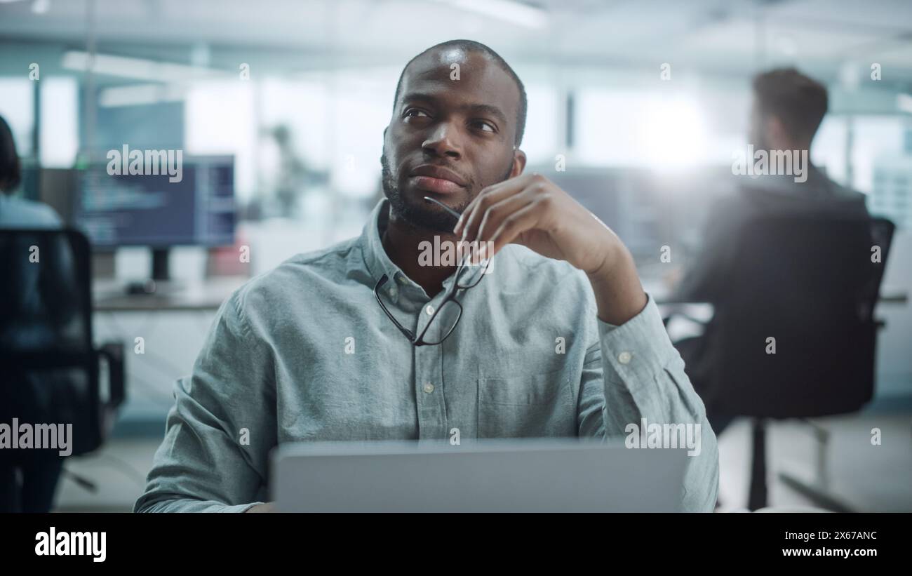 Modern Office: Portrait of Thoughtful Black IT Programmer Working on Laptop, Finding Solution, Solving Problems, Brainstorming. Male Software Engineer Wearing Glasses Develop App, Program, Video Game Stock Photo