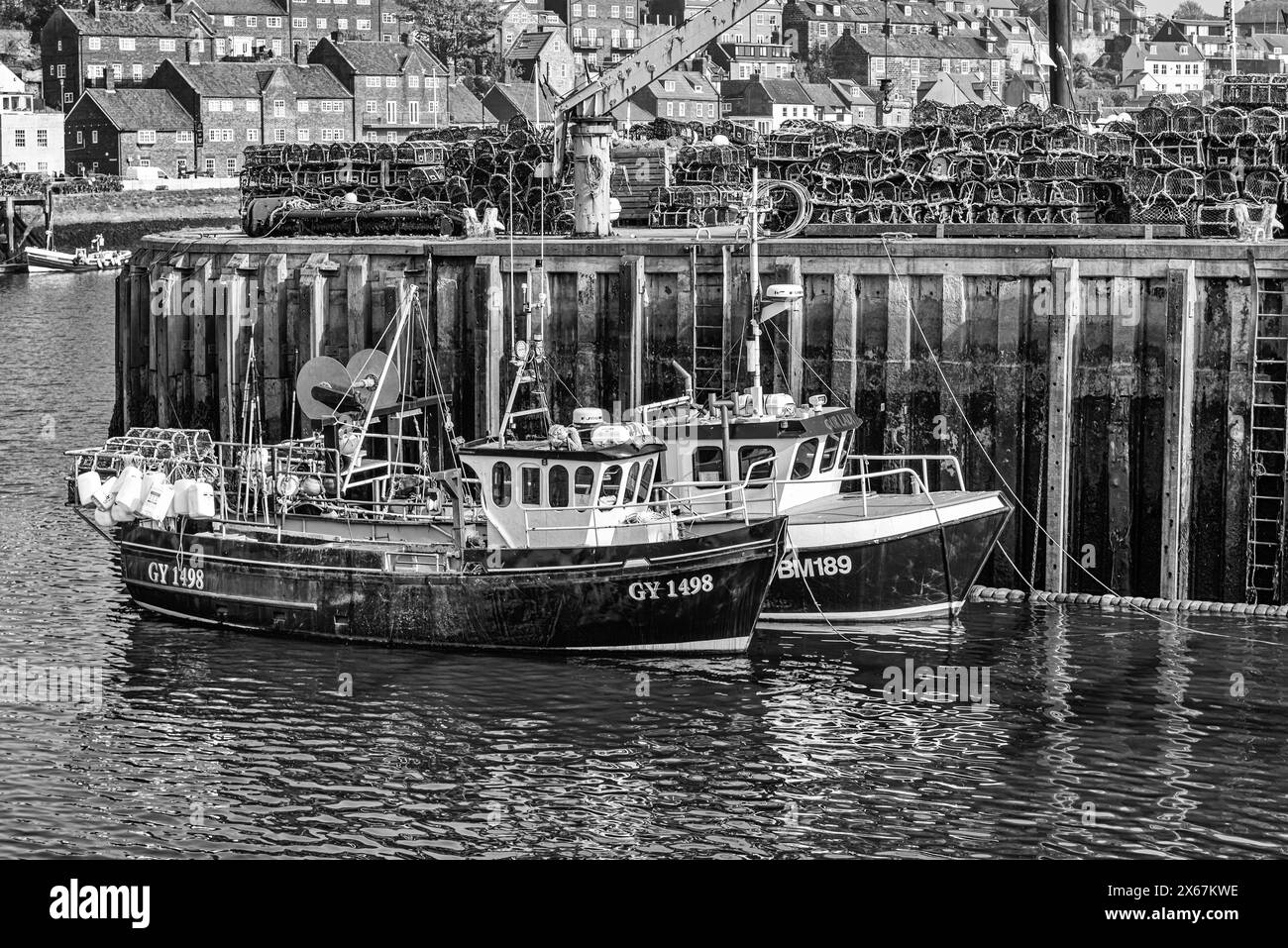 Two small fishing boats are moored alongside a quay and buildings are in the background. Lobster pots are stacked on the quayside. Stock Photo