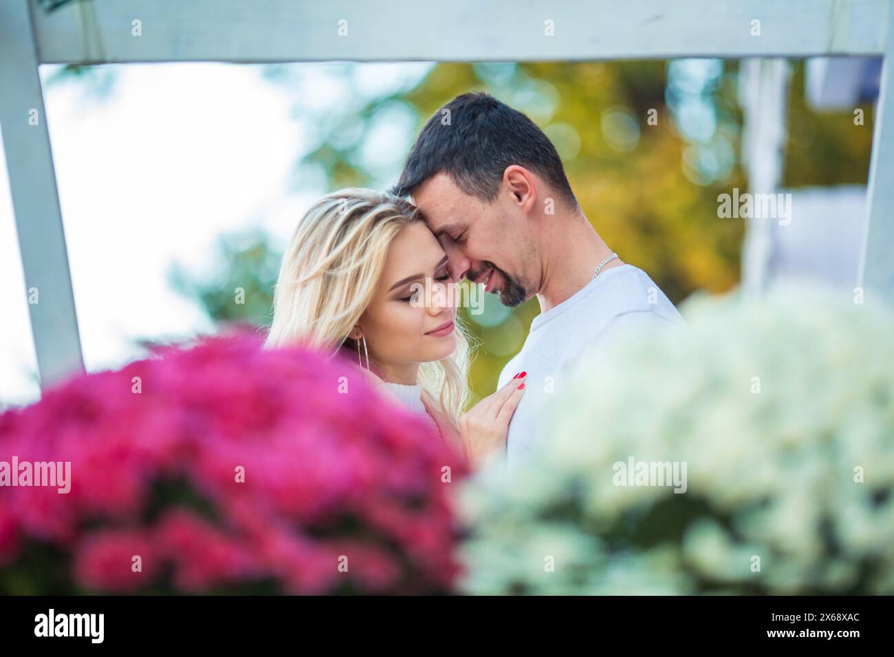Love story engagement session of a happy young caucasian straight couple in white sweaters, boyfriend is hugging girlfriend, flowers foreground, blurr Stock Photo
