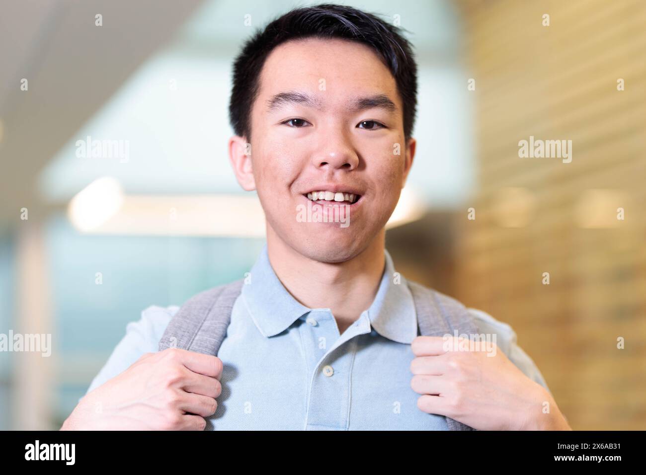 A young teenage Asian male college student is standing in a hallway with his backpack with him for school. He is grinning at the camera. Stock Photo