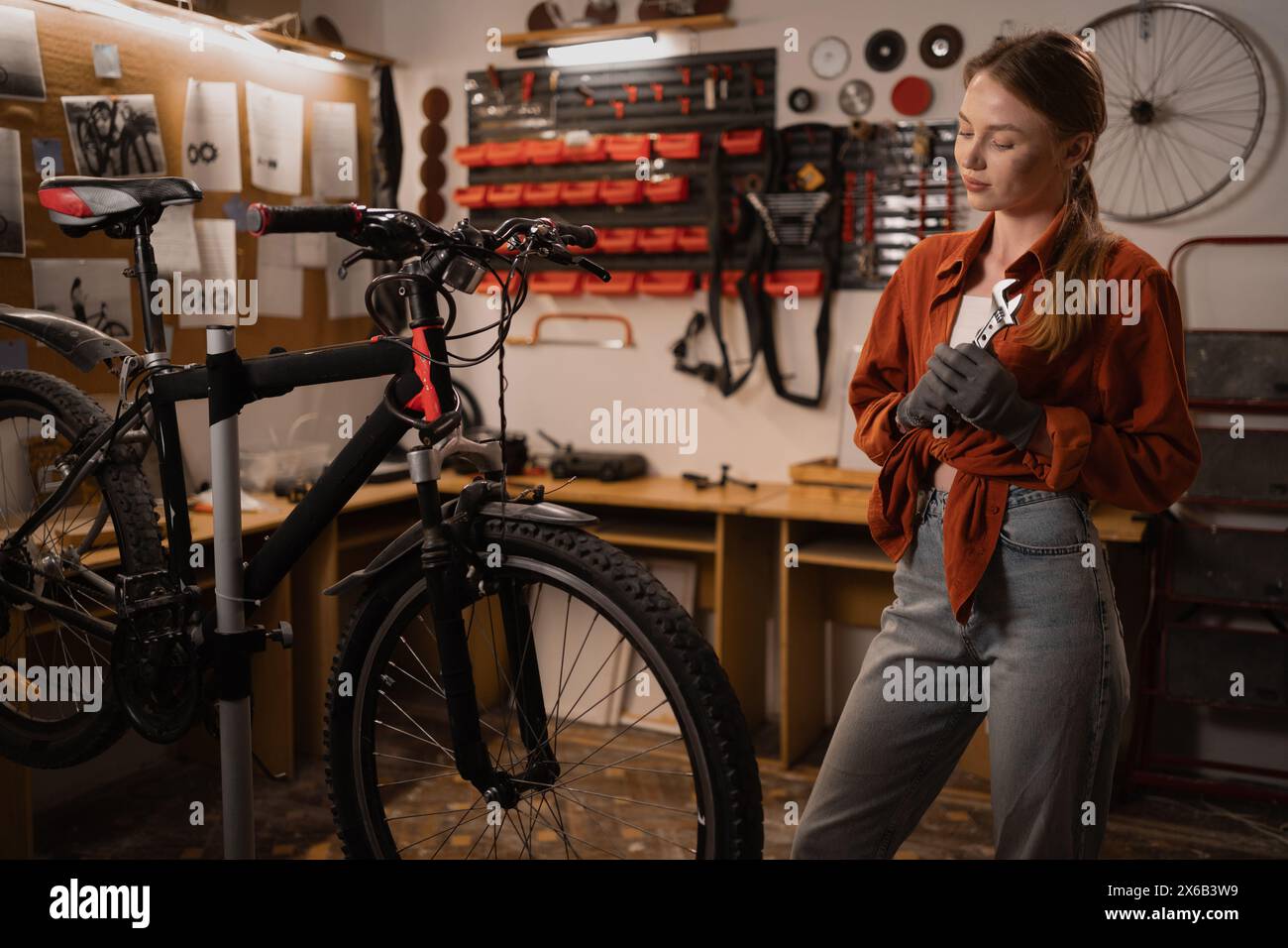 Portrait of a blonde girl standing with an adjustable wrench in her hands, repairing a bicycle in a workshop or garage. Hobby and DIY concept Stock Photo