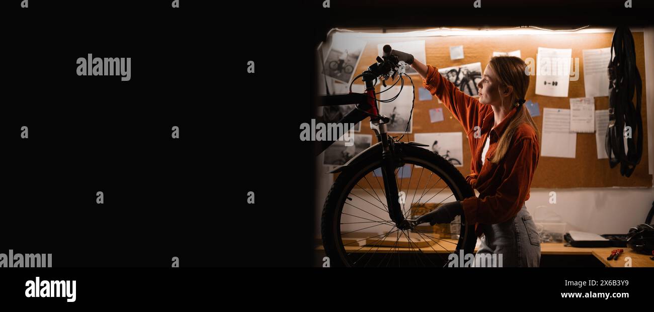 A young woman repairing a bicycle in an atmospheric garage, fixing the steering wheel after an accident. Hobby and DIY concept Stock Photo