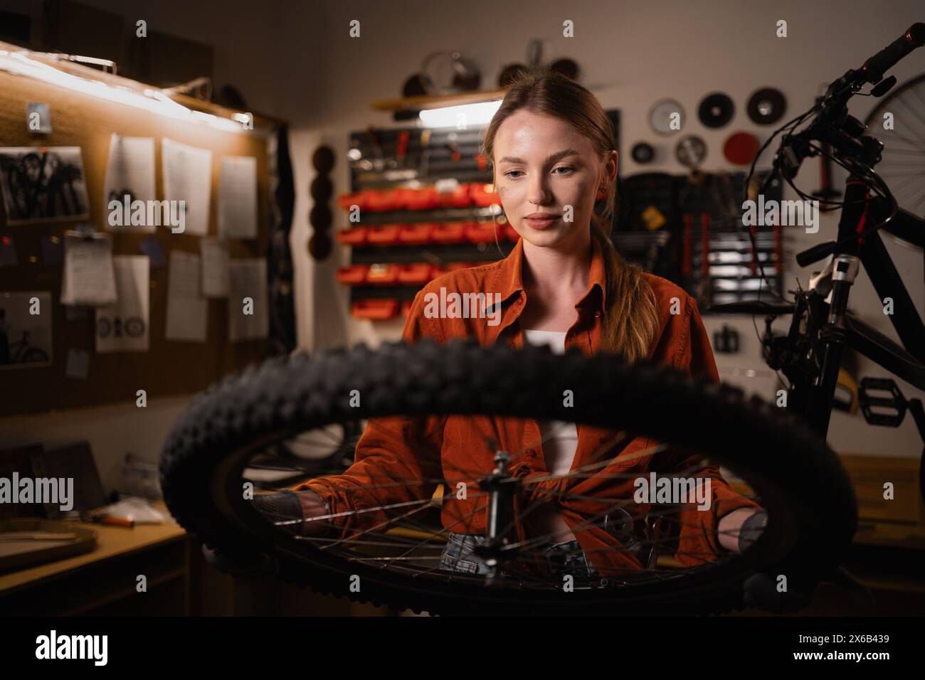 A young beautiful blonde woman holds a bicycle wheel while standing in a workshop or garage, doing her favorite hobby. Bicycle maintenance concept Stock Photo