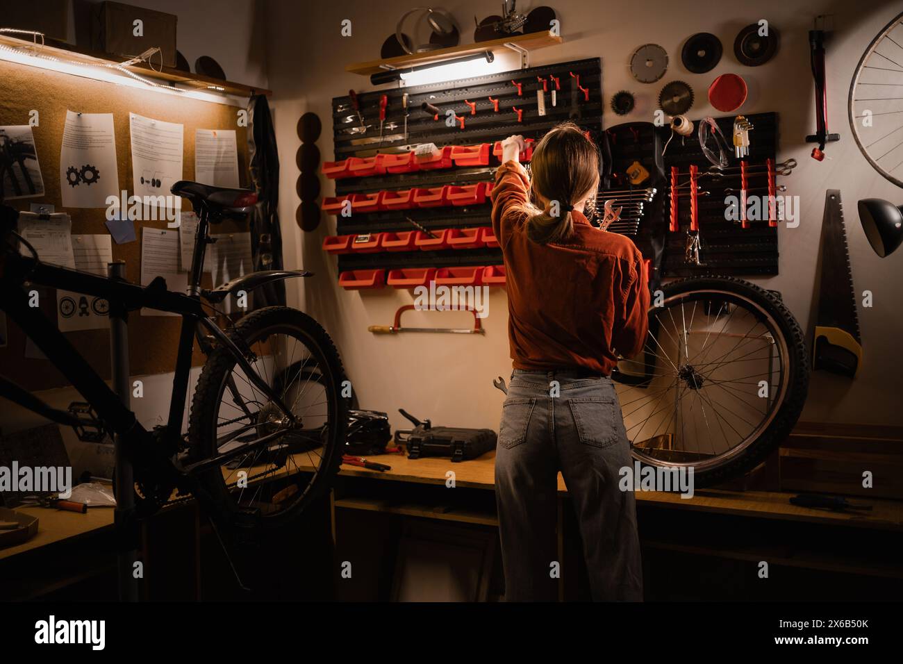 Rear view of young woman standing near wall with tools in workshop. Bicycle repair and maintenance concept Stock Photo