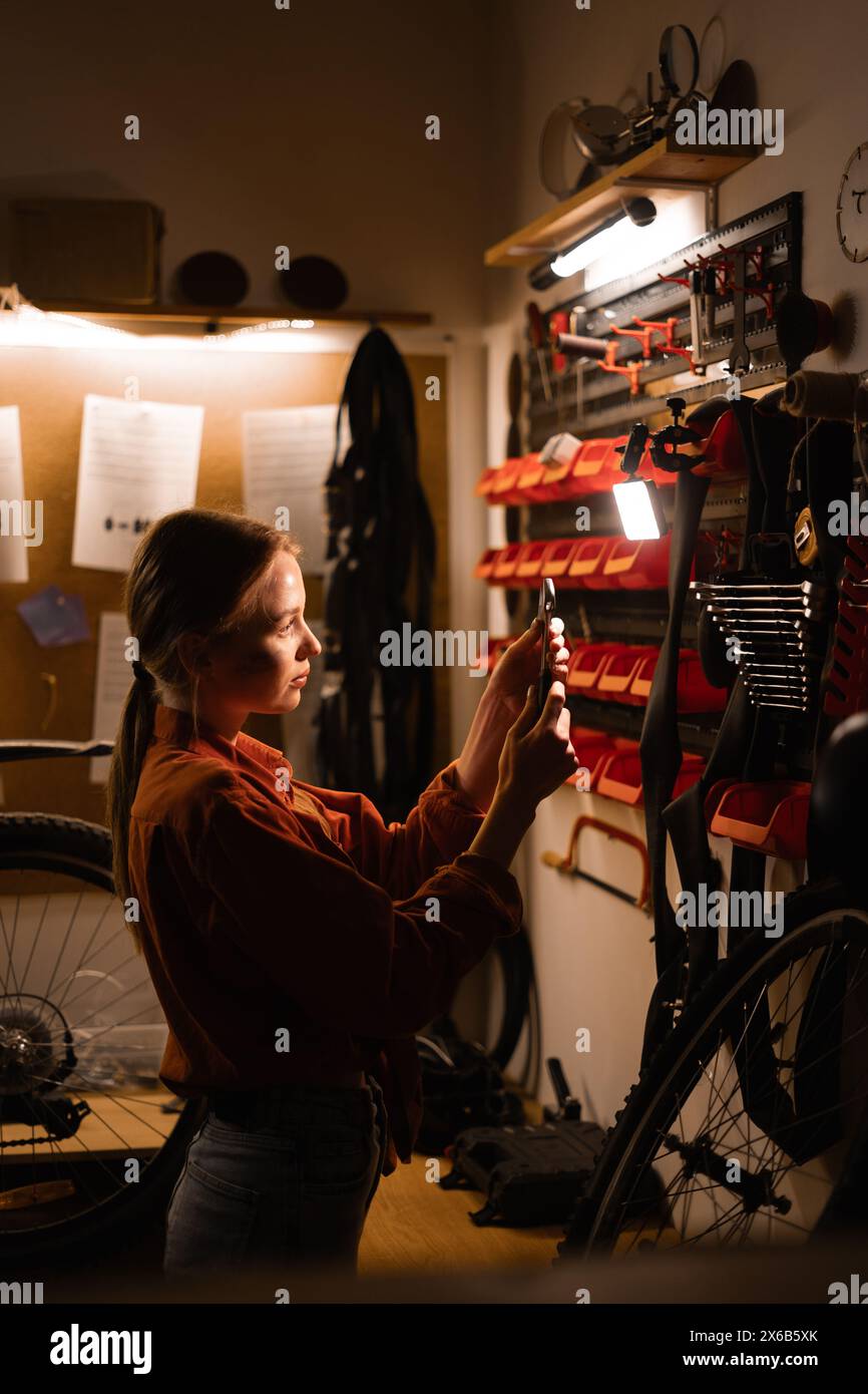 Female mechanic repairing bicycle in workshop, woman standing near wall with tools in garage. Bicycle maintenance concept. Stock Photo