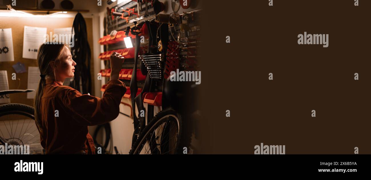 Female mechanic repairing bicycle in workshop, woman standing near wall with tools in garage. Bicycle maintenance concept Stock Photo