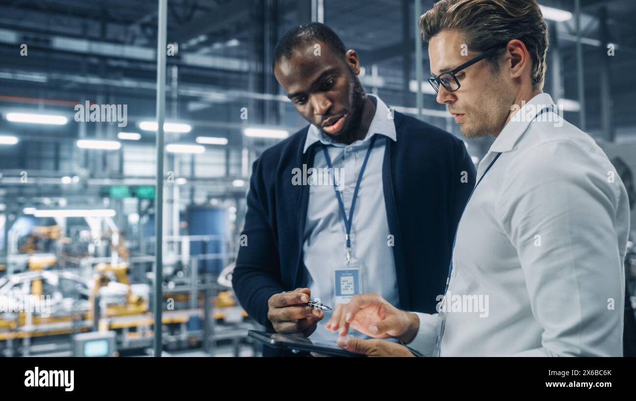 Two Diverse Automotive Industrial Engineers Talking About Vehicle Production while Standing in Office at a Car Assembly Plant. Industrial Specialists Discuss Work Projects on Tablet Computer. Stock Photo