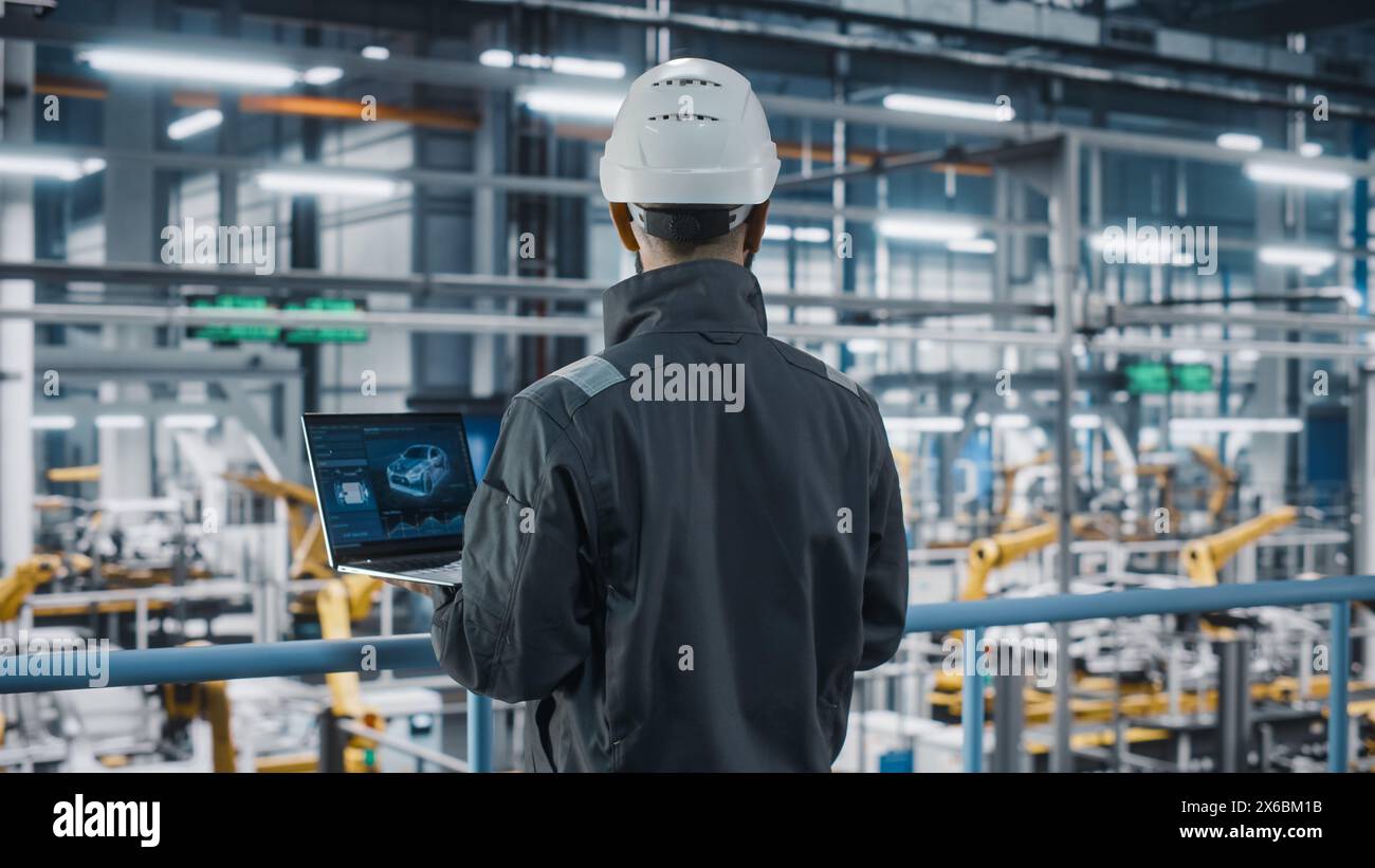 Car Factory Engineer in Work Uniform Using Laptop Computer. Automotive Industrial Manufacturing Facility Working on Vehicle Production with Robotic Arms Technology. Automated Assembly Plant. Stock Photo