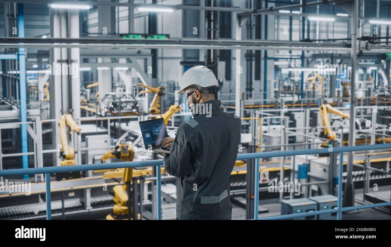 Car Factory Engineer in Work Uniform Using Laptop Computer. Automotive Industrial Manufacturing Facility Working on Vehicle Production with Robotic Arms Technology. Automated Assembly Plant. Stock Photo