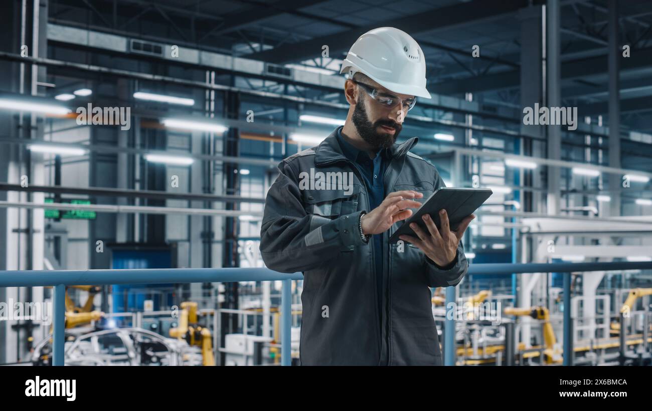 Portrait of Automotive Industry Engineer in Safety Glasses and Uniform Using Laptop at Car Factory Facility. Professional Assembly Plant Specialist Working on Manufacturing Modern Electric Vehicles. Stock Photo