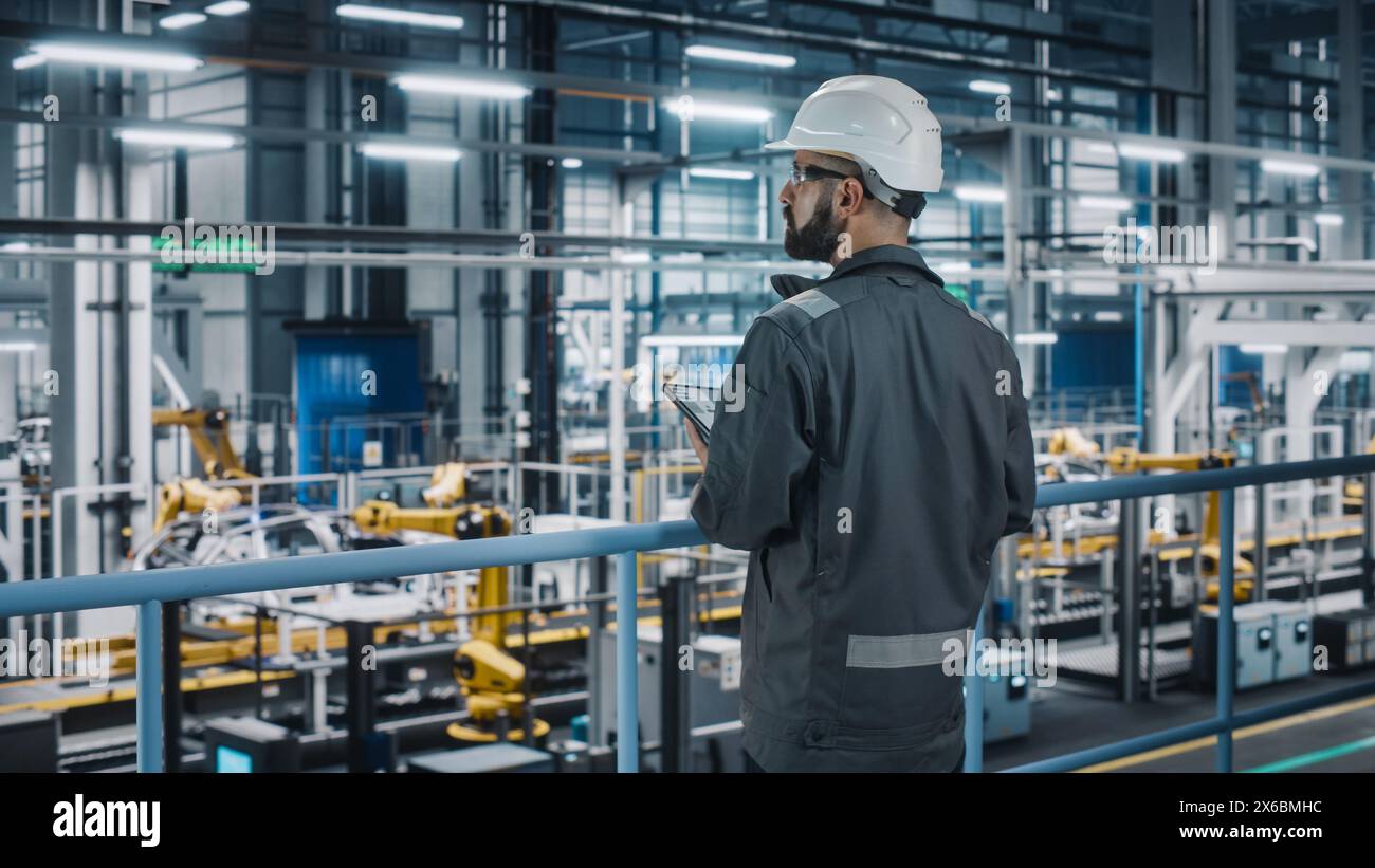 Car Factory Engineer in Work Uniform Using Tablet Computer. Automotive Industrial Manufacturing Facility Working on Vehicle Production with Robotic Arms Technology. Automated Assembly Plant. Stock Photo