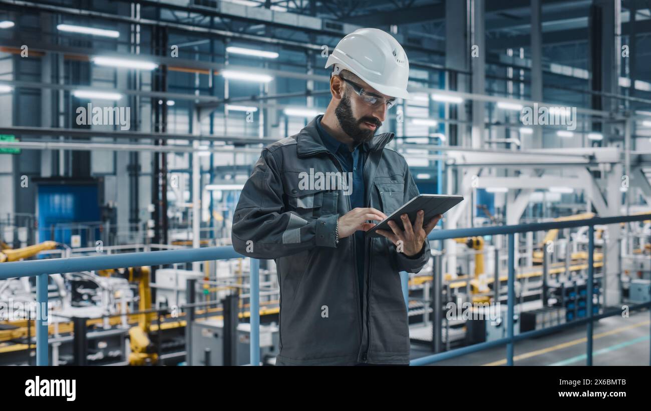 Portrait of Automotive Industry Engineer in Safety Glasses and Uniform Using Laptop at Car Factory Facility. Professional Assembly Plant Specialist Working on Manufacturing Modern Electric Vehicles. Stock Photo