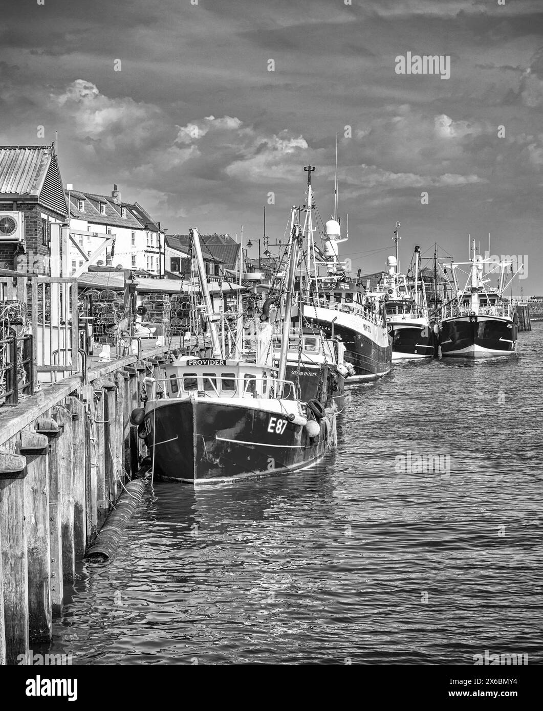 Whitby harbour with fishing boats moored alongside a wharf.  Buildings line the waterfront and a sky with clouds is above. Stock Photo