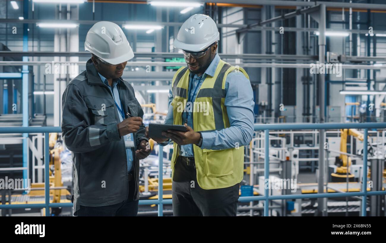 Multiethnic Manager and African American Car Factory Engineer in Uniform Using Tablet Computer. Automotive Industry 4.0 Manufacture Employees Discuss Work on Vehicle Assembly Plant. Stock Photo
