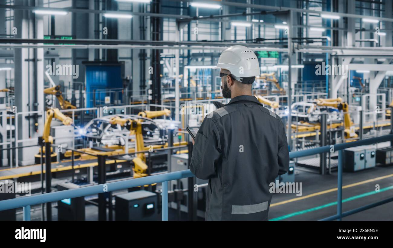 Car Factory Engineer in Work Uniform Using Tablet Computer. Automotive Industrial Manufacturing Facility Working on Vehicle Production with Robotic Arms Technology. Automated Assembly Plant. Stock Photo