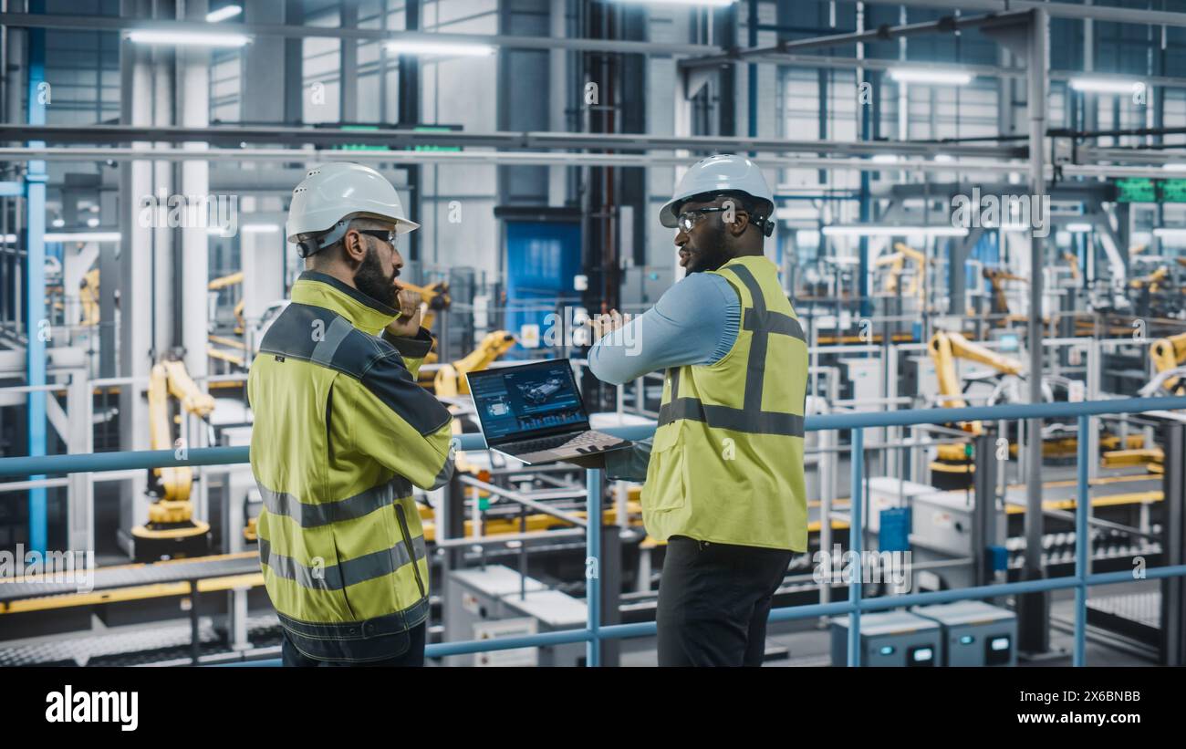 Two Car Factory Specialists in High Visibility Vests Using Laptop Computer. Engineers Discussing Automotive Industrial Manufacturing Facility Process of Vehicle Production. Automated Assembly Plant. Stock Photo