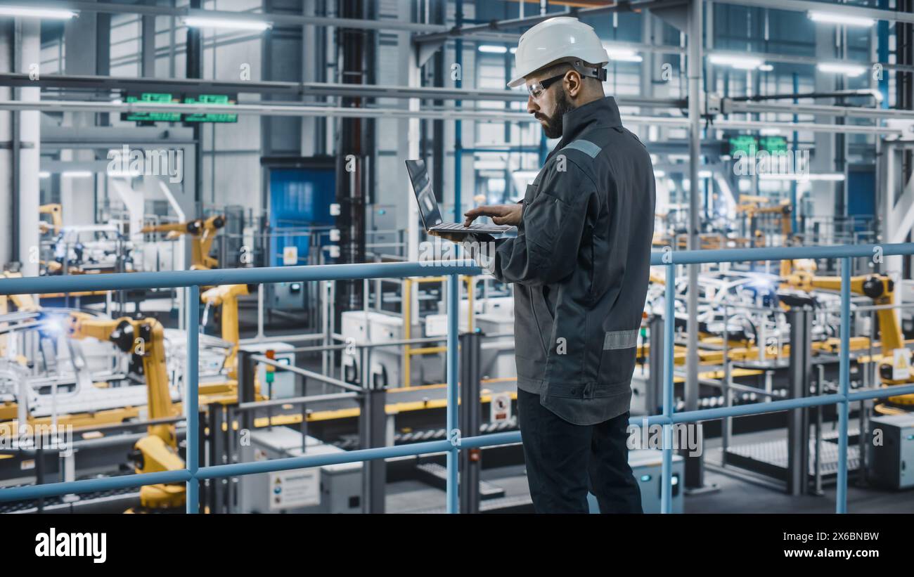 Car Factory Engineer in Work Uniform Using Laptop Computer. Automotive Industrial Manufacturing Facility Working on Vehicle Production with Robotic Arms Technology. Automated Assembly Plant. Stock Photo