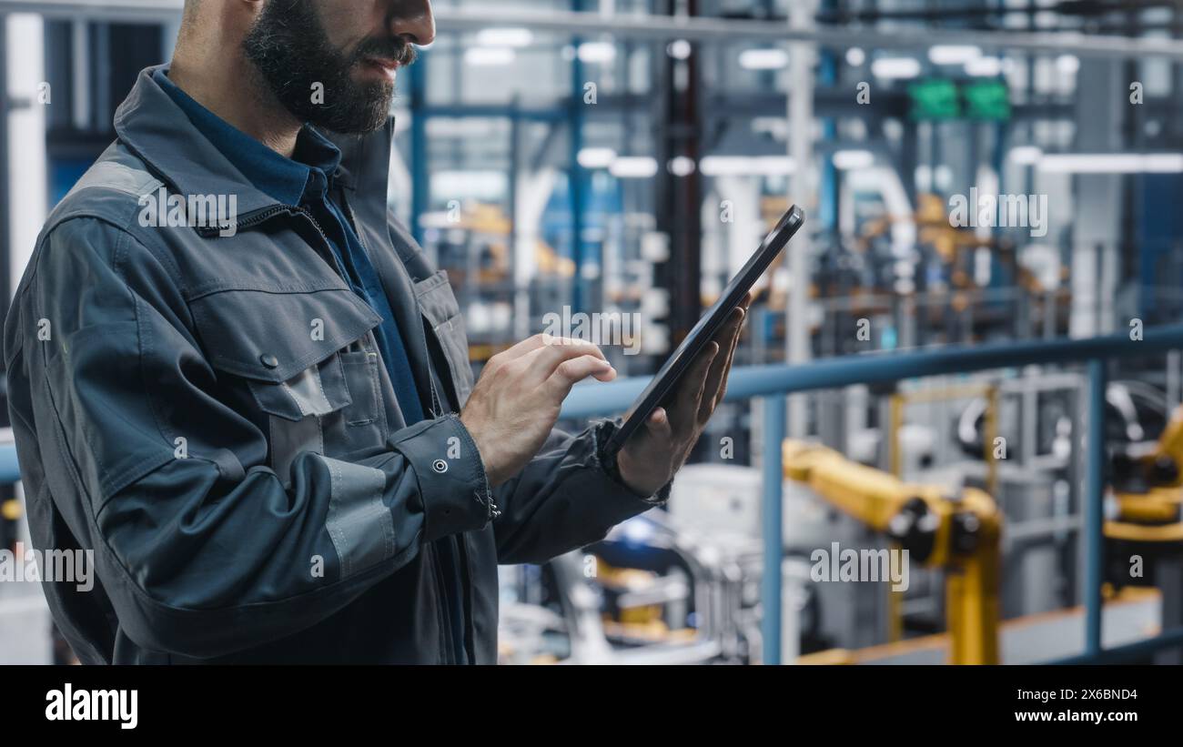 Portrait of Automotive Industry Engineer in Safety Glasses and Uniform Using Tablet at Car Factory Facility. Professional Assembly Plant Specialist Working on Manufacturing Modern Electric Vehicles. Stock Photo