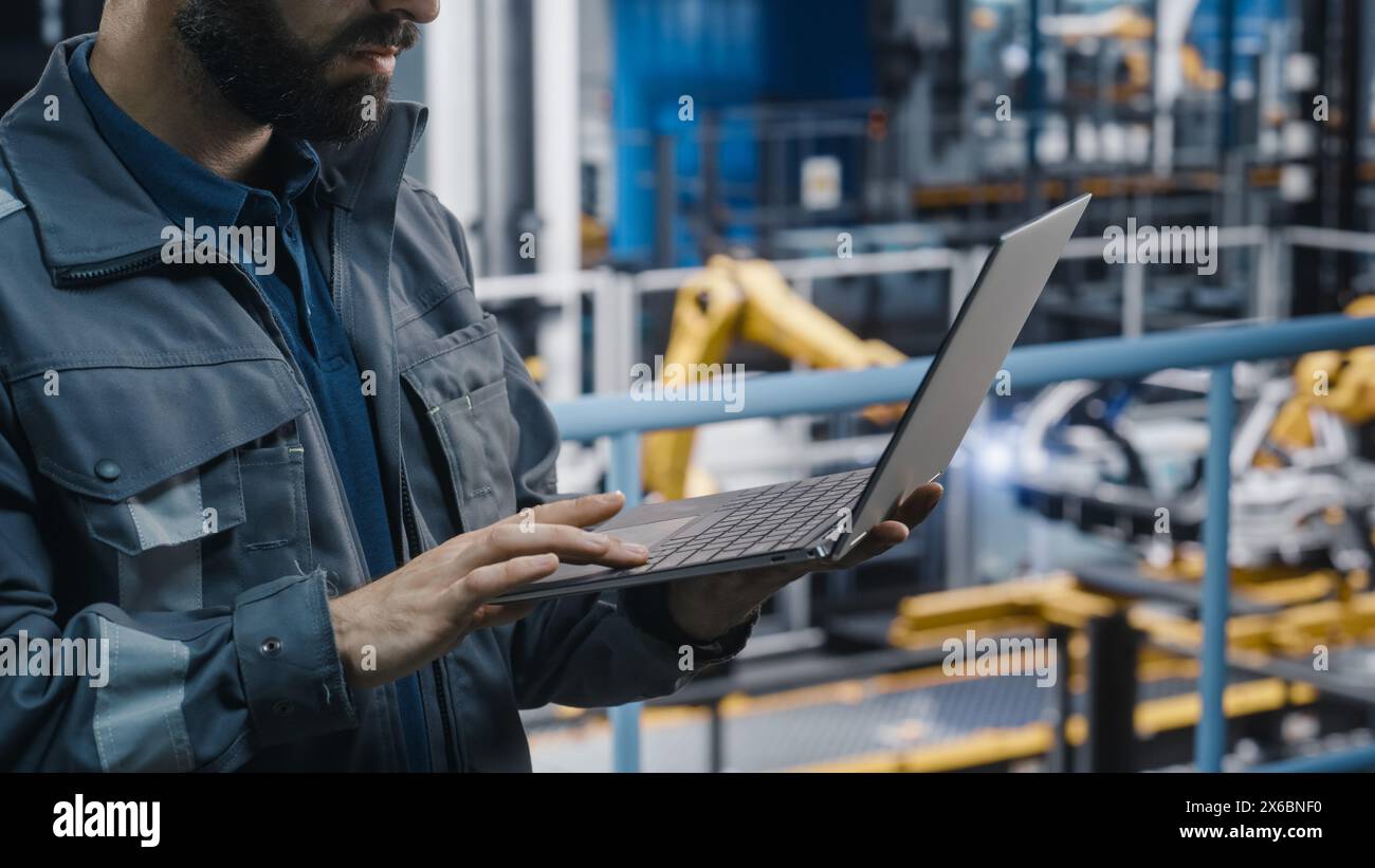 Close Up of Automotive Industry Engineer in Grey Uniform Using Laptop at Car Factory Facility. Professional Assembly Plant Specialist Working on Manufacturing Modern Electric Vehicles. Stock Photo