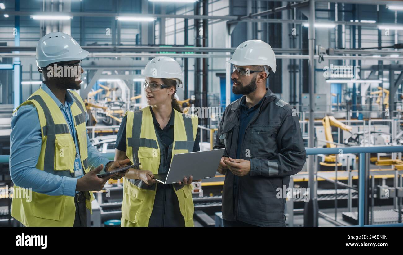 Young Diverse Team of Car Factory Specialists Working on Laptop and Tablet Computers. Engineers Discussing Automotive Industrial Manufacturing Technology on Modern Vehicle Assembly Plant. Stock Photo