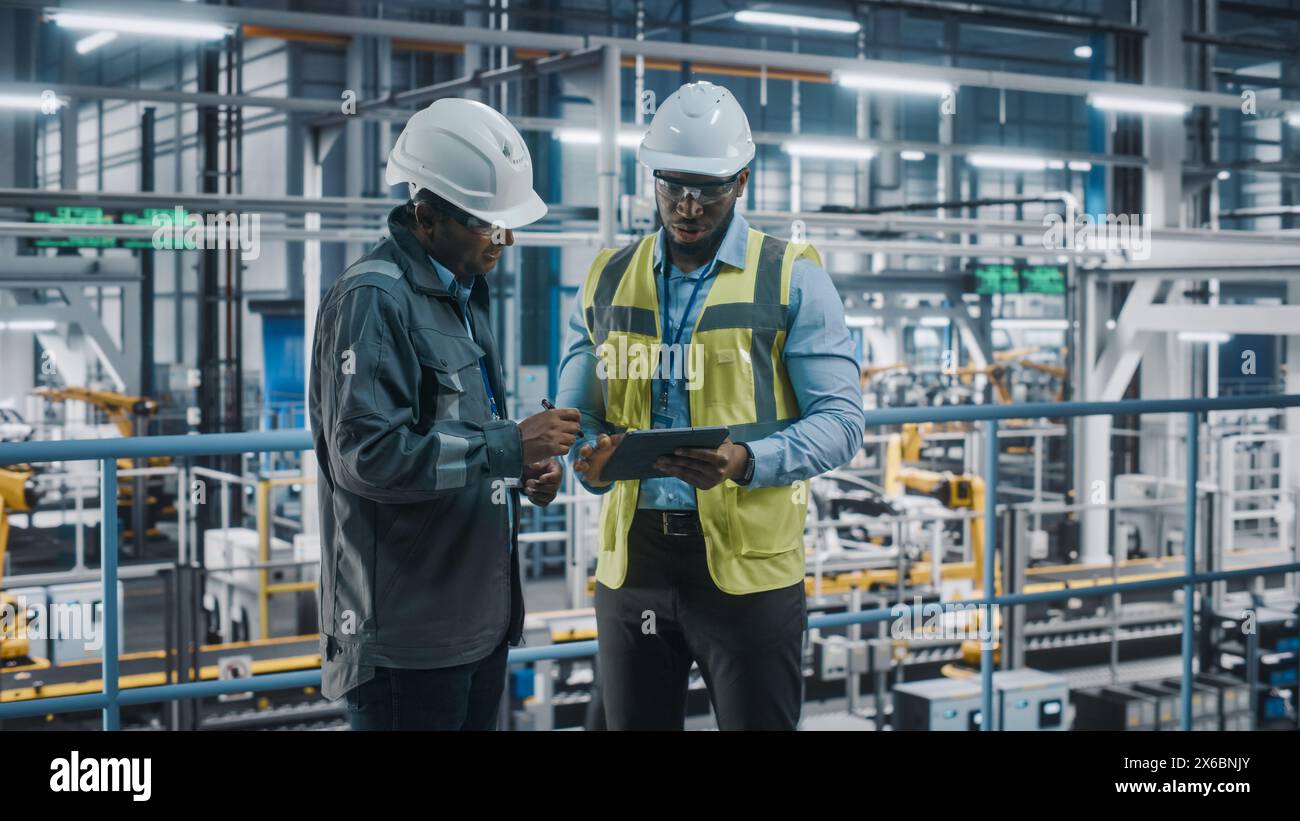 Multiethnic Manager and African American Car Factory Engineer in Uniform Using Tablet Computer. Automotive Industry 4.0 Manufacture Employees Discuss Work on Vehicle Assembly Plant. Stock Photo