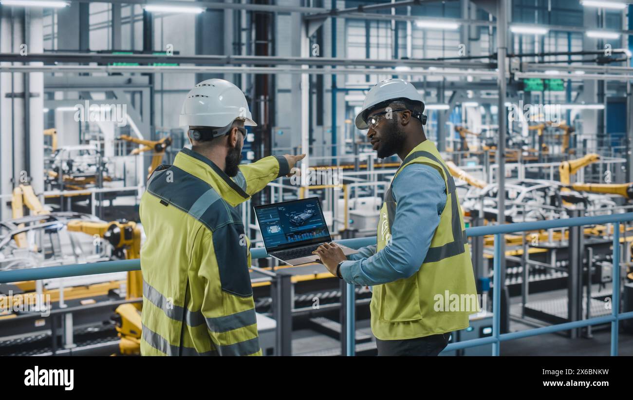 Two Car Factory Specialists in High Visibility Vests Using Laptop Computer. Engineers Discussing Automotive Industrial Manufacturing Facility Process of Vehicle Production. Automated Assembly Plant. Stock Photo