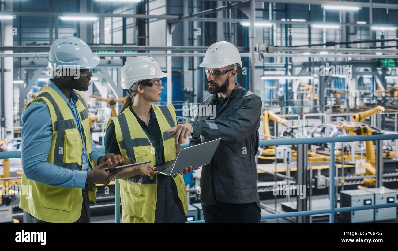 Young Diverse Team of Car Factory Specialists Working on Laptop and Tablet Computers. Engineers Discussing Automotive Industrial Manufacturing Technology on Modern Vehicle Assembly Plant. Stock Photo