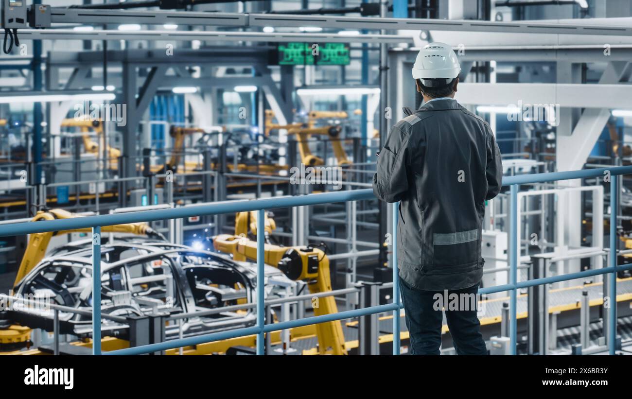 Multiethnic Car Factory Engineer in Work Uniform Using Tablet Computer. Automotive Industrial Manufacturing Facility Working on Vehicle Production with Robotic Arms. Automated Assembly Plant. Stock Photo