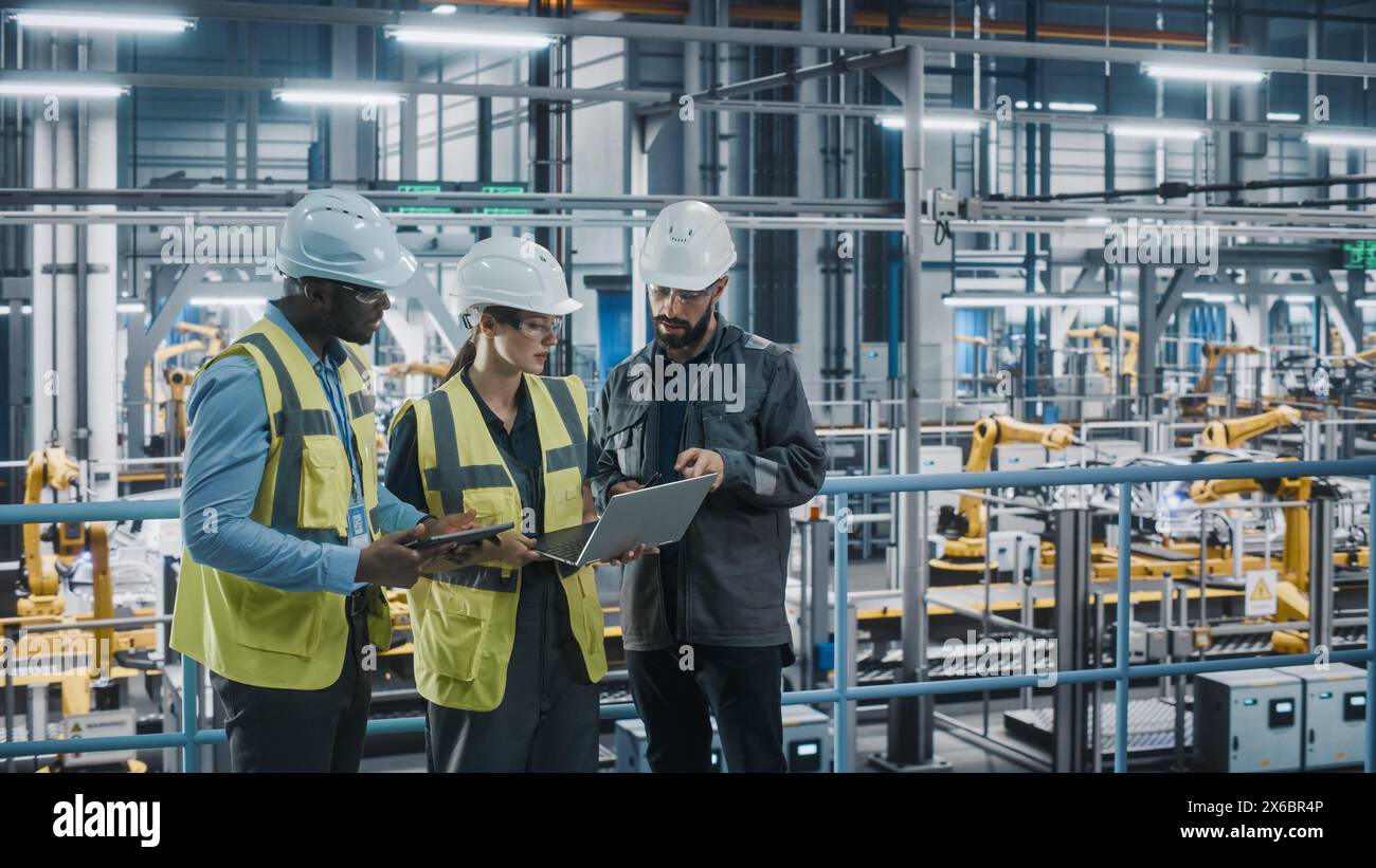 Young Diverse Team of Car Factory Specialists Working on Laptop and Tablet Computers. Engineers Discussing Automotive Industrial Manufacturing Technology on Modern Vehicle Assembly Plant. Stock Photo