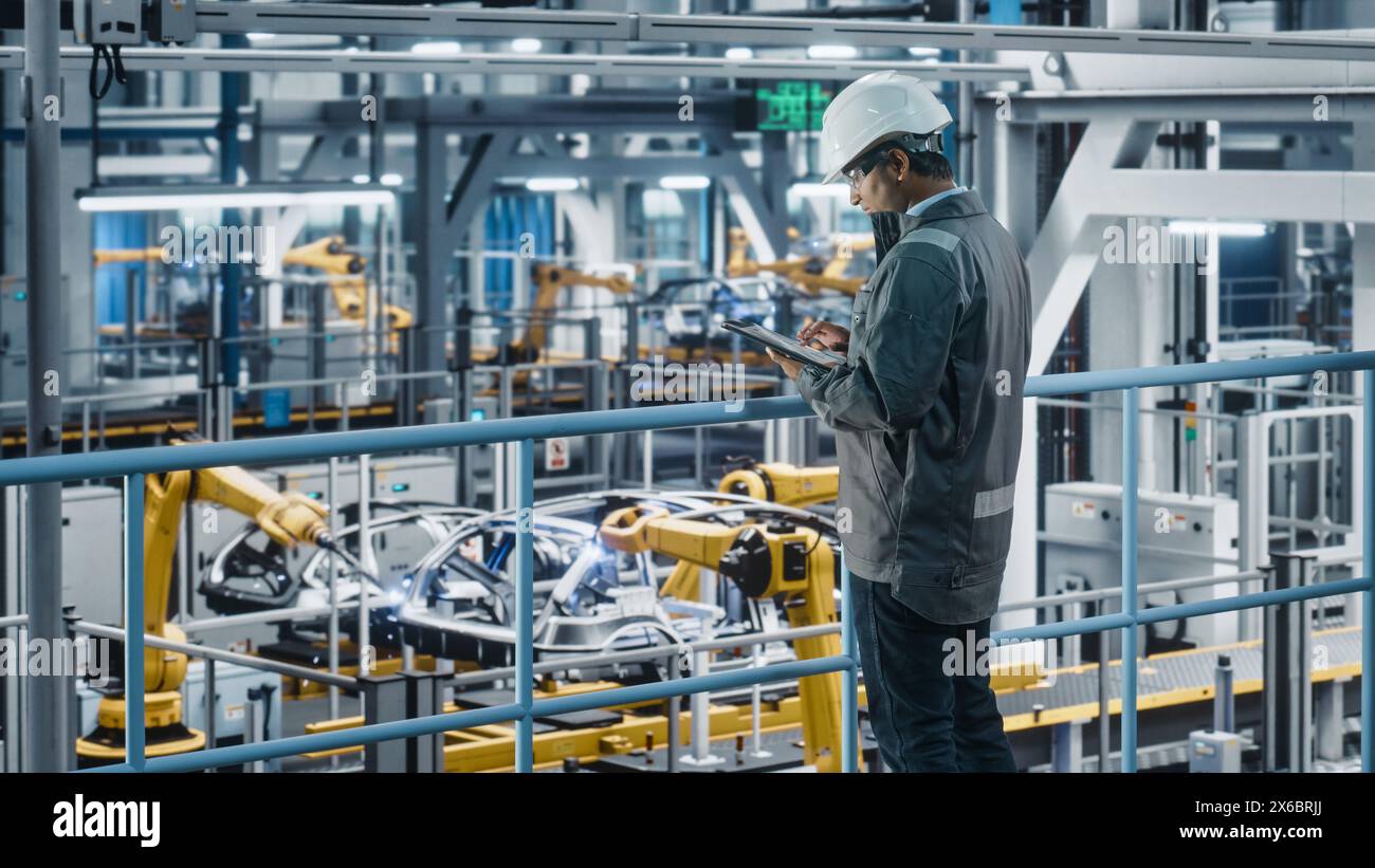 Multiethnic Car Factory Engineer in Work Uniform Using Tablet Computer. Automotive Industrial Manufacturing Facility Working on Vehicle Production with Robotic Arms. Automated Assembly Plant. Stock Photo
